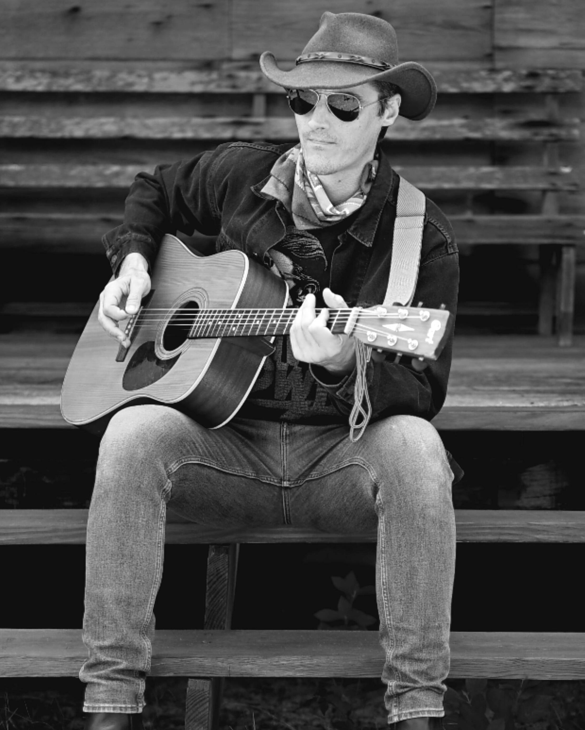 A man dressed as a cowboy sits on a rustic porch playing a guitar. Photo by Dylan Jon Wade Cox.